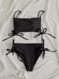 Flat lay of black two-piece bikini with side tie details on soft white fabric background