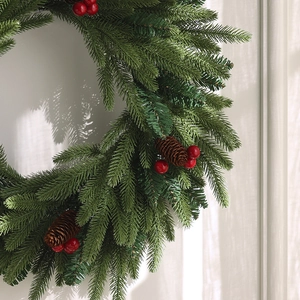 Christmas wreath with green pine needles, red berries and pine cones on white wall