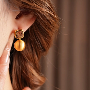 Close-up of golden geometric earrings with textured square stud and smooth sphere dangle