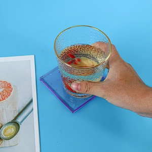 Detail showing a hand holding a gold-rimmed glass on a purple acrylic coaster with blue background