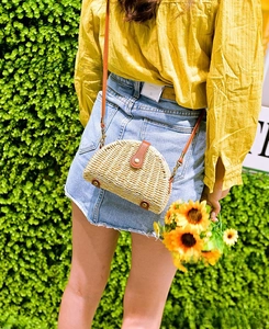 Outdoor shot of semicircular straw crossbody bag with brown leather strap and metal studs