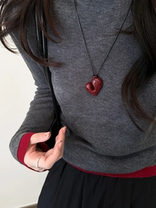 Dettaglio di un ciondolo a cuore rosso su cordino nero indossato su maglia grigia