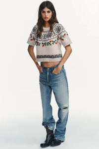 Detail of a beige knit crop top with red floral embroidery and distressed blue jeans