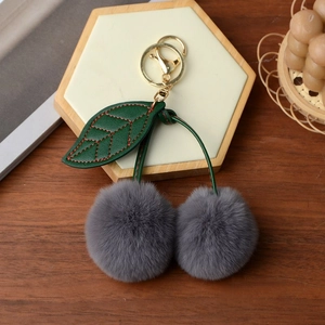 Detail of grey plush cherry keychain with soft faux fur balls and green leaf on wooden desk