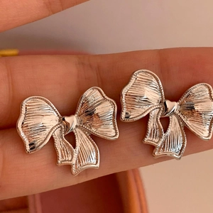 Silver bow stud earrings with pleated texture and tiny heart detail shown on finger
