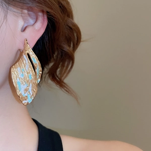 Detail of leaf-shaped gold stud earring with blue-green glaze worn on model's ear