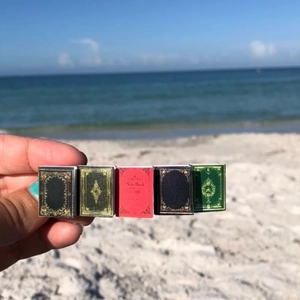 Close-up of five colorful miniature book hair clips held against a sunny beach background