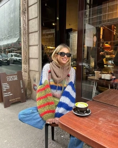 Detail shot of a relaxed rainbow striped knit sweater styled with a scarf in a street cafe