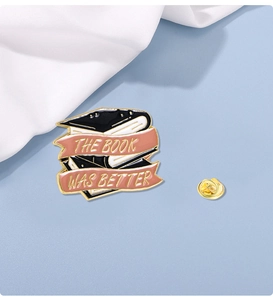 Close-up of enamel book brooch with pink banner reading THE BOOK WAS BETTER in gold finish