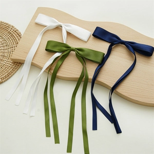 Three long satin bow hair clips in white, green, and navy blue on a wooden tray