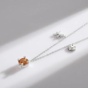 Silver necklace with orange gemstone and bow charm displayed with minimalist diagonal shadows