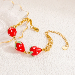 Detail of a golden double-layered bracelet featuring three red strawberry charms and heart tag