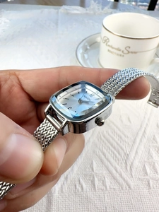 Elegant woman holding a square silver watch with mesh band over a lace setting