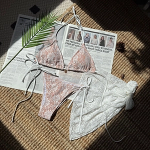 Flat lay of white floral lace three-piece bikini set with sheer mesh skirt and beach props