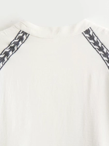 Detailed close-up of ethnic contrast embroidery on white raglan shoulder blouse fabric
