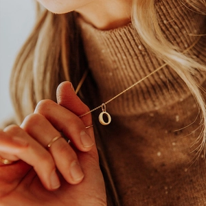 Macro detail of a gold-colored letter O pendant necklace worn over a warm brown sweater.