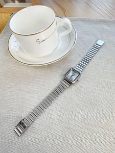 Silver square dial metal watch with chain strap displayed on beige textured fabric