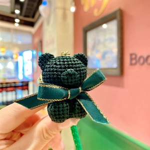 Macro detail of dark green woven bear keychain featuring a luxury satin bow with gold trim