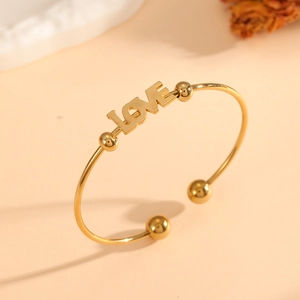 Minimalist gold LOVE engraved open bangle with round bead accents on a warm beige background
