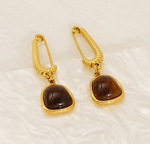 Detailed view of gold safety pin earrings with square dark gemstone pendants on white fabric
