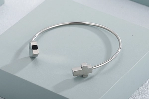 Close-up of silver open cuff bracelet featuring heart and cross charms on a blue pedestal