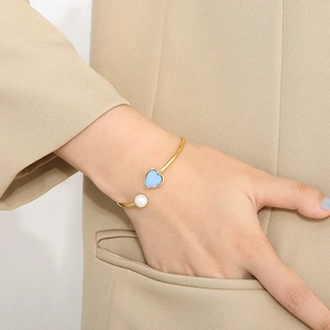 Detailed close-up of golden open bangle with blue heart and white pearl on wrist