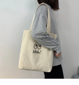 Detalle del bolso tote de lona beige con diseño de ojos y texto Shh! en fondo minimalista