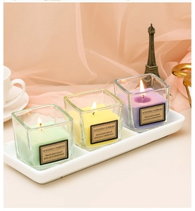 Close-up of pastel scented candles in square glass jars on a white ceramic tray