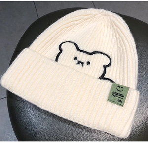Detailed view of cream knit beanie featuring bear embroidery and green smiley face label