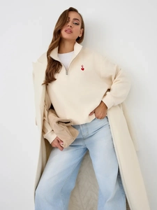 Close-up of minimalist heart embroidery on beige half-zip sweatshirt and light blue jeans