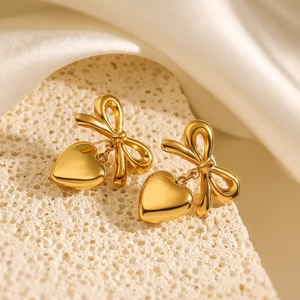 Golden bow heart earrings made of stainless steel displayed on beige stone and silk
