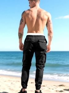 Back view of men's black cargo pants with multiple pockets and elastic cuffs in outdoor beach setting