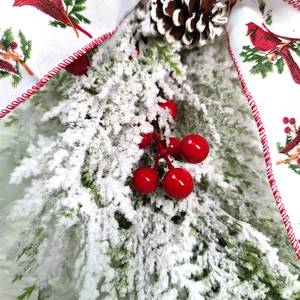 Close-up of snowy pine needles, red berries, and cones with a cardinal ribbon detail