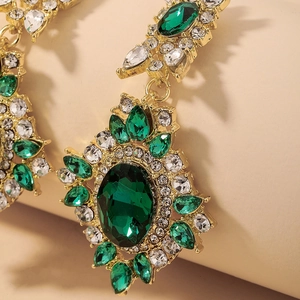 Macro detail of gold flower earrings featuring a large emerald green oval gem and rhinestones