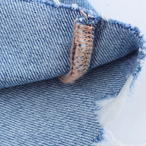 Close-up of light blue denim fabric featuring orange stitching and frayed edge detail