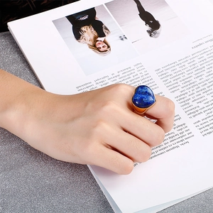 Golden ring with heart-shaped blue stone worn over a magazine on shiny silver background