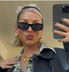 Selfie view of woman wearing punk oval sunglasses with chunky gold earrings and necklace