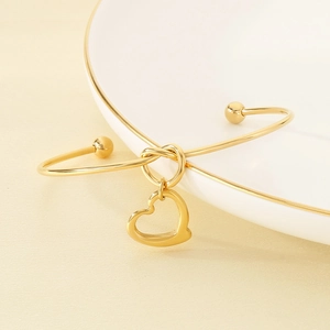 Detail view of a 18K gold-plated knotted bangle with a hollow heart pendant on a white ceramic plate
