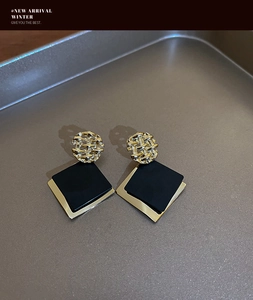 Detail of square black drop earrings with golden braided texture on a grey tray