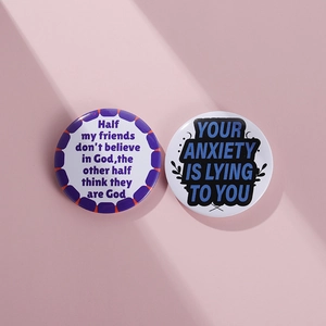 Close-up of two round tinplate badges with inspirational quotes on a soft pink surface