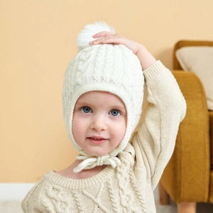 Close-up of baby white cable knit hat featuring a pompom and cozy earflaps