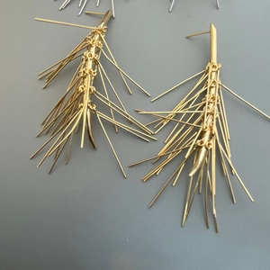 Dettaglio di orecchini ad ago di pino dorati con lunghe frange metalliche su grigio