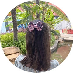 Pink sequin bow hair clip worn by a girl outdoors with palm trees and a pond background