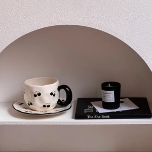 Irregular ceramic coffee cup and saucer set with black cherry pattern in a cozy shelf setting