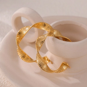 Golden twisted hoop earrings with textured hollow pattern on white ceramic stand