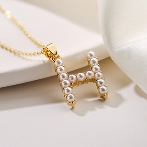 Elegante collana dorata con ciondolo a lettera H e perle su tessuto bianco morbido