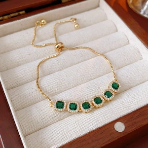 Detail of gold bracelet with square emerald green stones and white zircon halo in a wood box