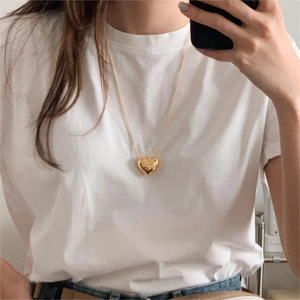 Mirror selfie showing a golden 3D heart pendant necklace worn over a white t-shirt
