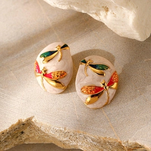 Macro detail of oval gold-plated earrings with colorful enamel dragonfly on marble base