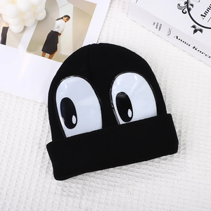 Detail view of a black knitted beanie featuring large cartoon eyes on white fabric background